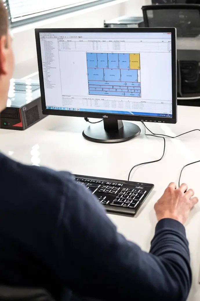 Imagem de um homem em frente a um computador. Na tela do computador, o software Ardis Optimizer aberto com o desenho de uma chapa de madeira preenchida com diversas peças de tamanho variado.