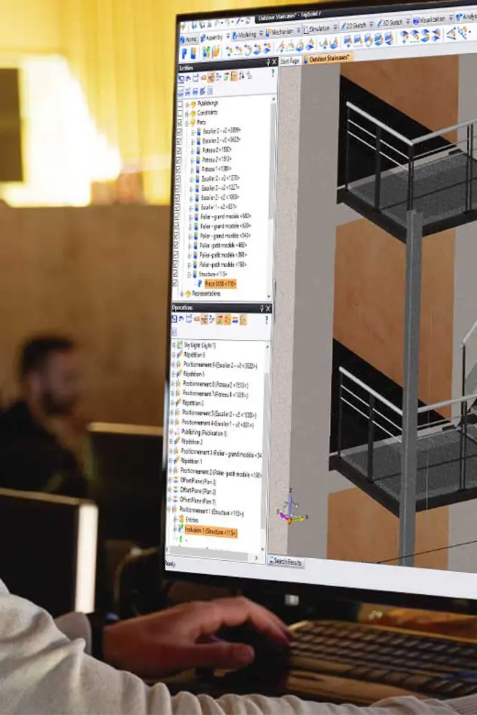 Imagem de um homem em frente a um computador. Na tela, um projeto do TopSolid de escadas metálicas em um prédio.